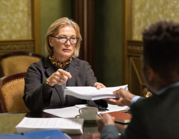 An older woman sits in a business meeting in a luxurious office. She is surrounded by papers.