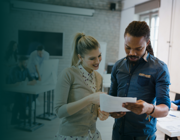 A man and a woman stand together looking at a piece of paper. They are in a busy meeting room with other people in the background. The man is black and the woman is white. They both wear professional clothing.