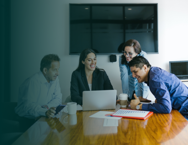 A team of coworkers including two men and two women sit at a table, looking at a laptop.