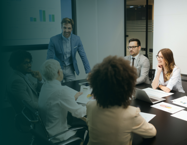 A team of professionals in a meeting. A young man in a blue suit stands at the end of a table in front of a business presentation.